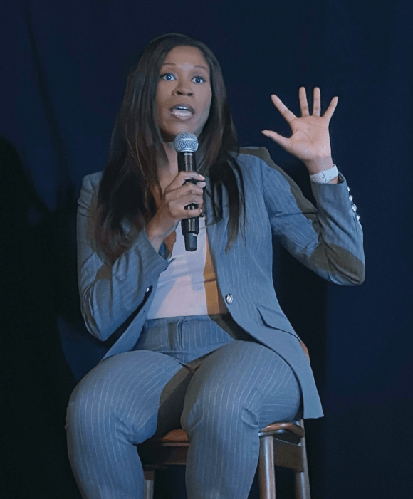 Julia Rock engages the audience while seated, wearing a pinstripe suit and holding a microphone mid-discussion under a spotlight.
